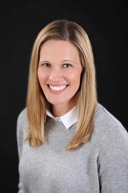 Professional woman with straight blonde hair smiles in a gray sweater and white collared shirt against a dark backdrop during her personal branding session in La Crosse, WI.