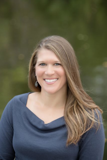 Professional woman with long hair and a navy top smiles in front of a blurred natural background during her personal branding portrait session in La Crosse, WI.