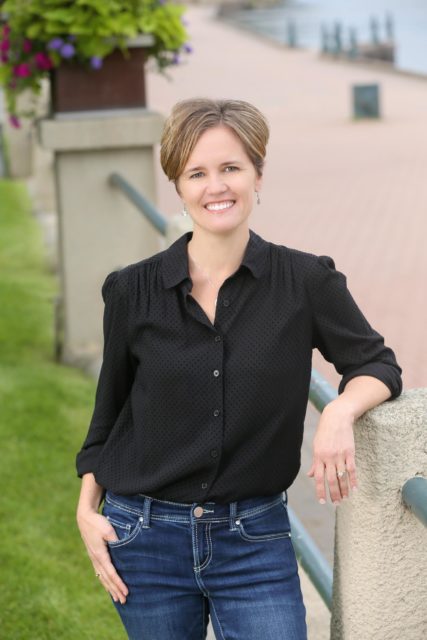 Professional woman in a black blouse and jeans smiles while leaning casually on a riverside railing, with flower planters and a brick path in the background in La Crosse, WI.
