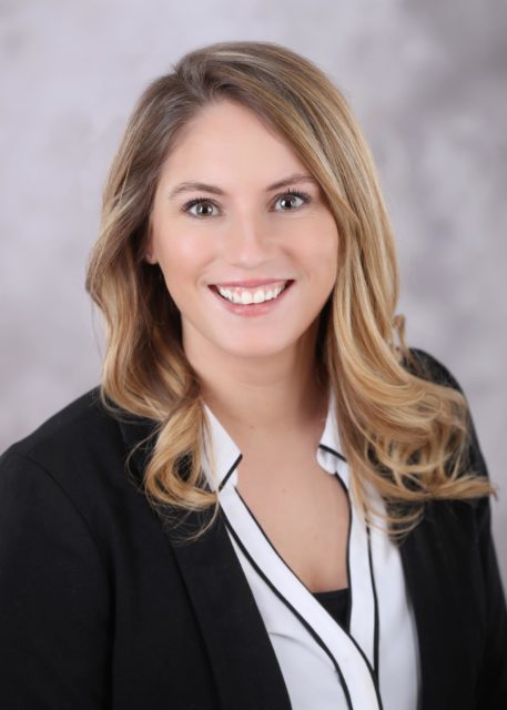 Professional woman with wavy blonde hair wears a black blazer over a white blouse with black trim, smiling confidently against a soft gray backdrop during her personal branding session in La Crosse, WI.